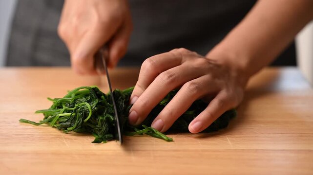 Woman hands using a knife to cut blanched spinach on a wooden cutting board, preparing ingredients for Korean spinach salad (Sigeumchi namul)