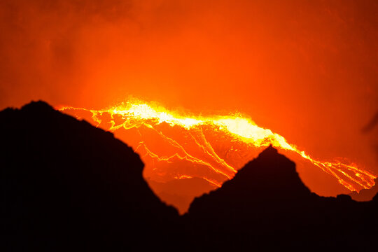 View of glowing lava flowing from the crater of Erta Ale volcano at night with dark volcanic silhouettes in the foreground, Ethiopia.