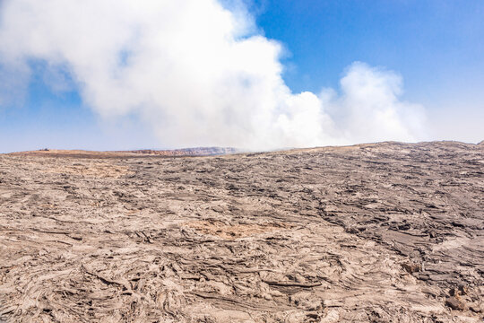View of the volcanic landscape of Erta Ale with solidified lava flows and volcanic smoke rising into a clear blue sky, Ethiopia.