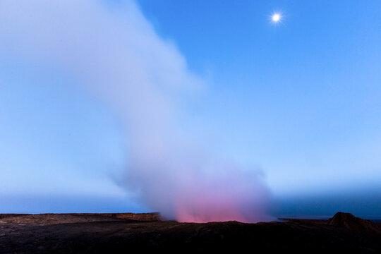 View of the glowing lava lake and volcanic smoke rising from the crater of Erta Ale volcano under a clear night sky.