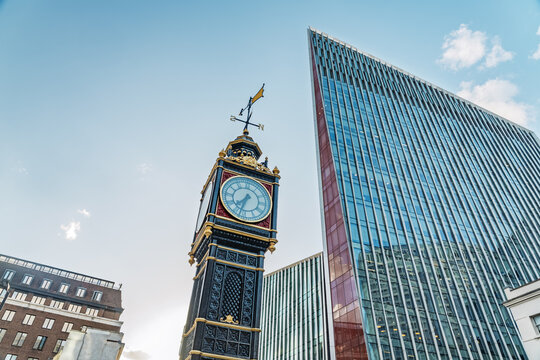 View of the Little Ben clock tower standing in contrast with modern glass skyscrapers under a clear blue sky in London, England, United Kingdom.