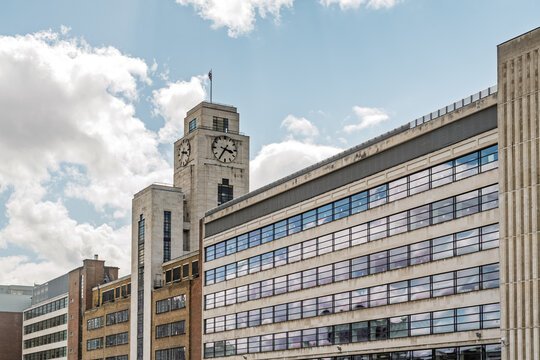 View of the iconic clock tower of Shell Mex House with its large clock face and stone facade, set against a bright blue sky with scattered white clouds in London, England, United Kingdom.