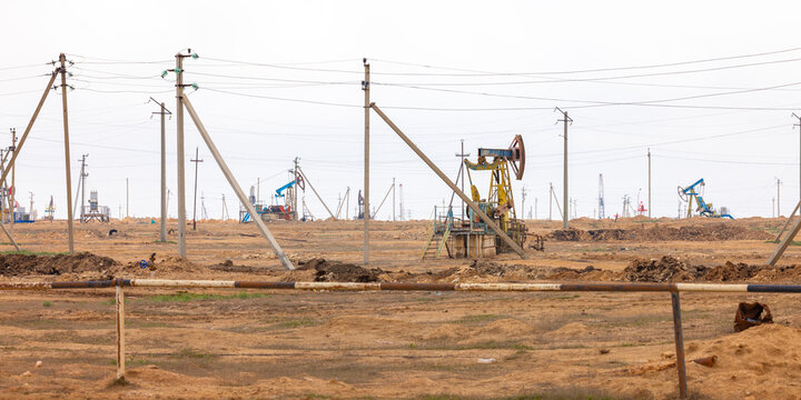 View of an oil field with pumpjacks operating in an arid landscape under an overcast sky, with power lines stretching across the horizon in Zhanaozen, Mangystau Region, Kazakhstan.