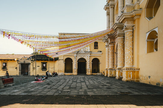 View of the ornate yellow facade of La Merced Church with decorative streamers hanging over the cobblestone plaza in Antigua Guatemala, Sacatepequez Department, Guatemala.