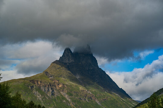View of the dramatic Otertinden mountain peak partially shrouded in heavy dark clouds above the rugged green slopes of the landscape in Storfjord, Troms, Norway.