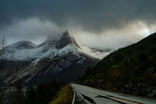 View of the snow-capped Stetind mountain peak under dramatic dark clouds with a road leading through the landscape in Narvik Municipality, Nordland, Norway.