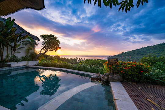 View of The Griya Villas and Spa featuring an infinity pool overlooking the ocean at sunset with lush tropical greenery and a dramatic cloudy sky in Kecamatan Abang, Bali, Indonesia.