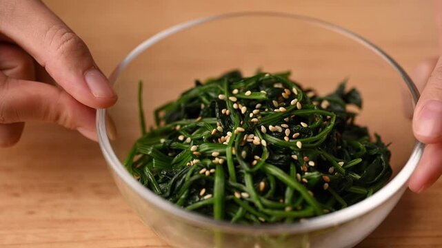 Close-up of hands rotating a bowl with Korean spinach salad (Segeumchi namul) on wooden table