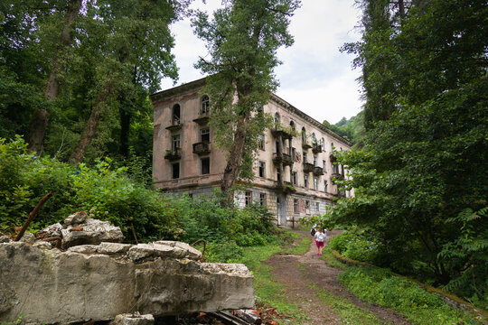 Abandoned multi-story building in an overgrown lush forest, Akarmara, Abkhazia. Tourist with a backpack walking on a dirt path towards a decaying historic structure.