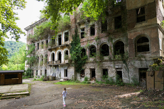 Young girl walking towards a massive overgrown abandoned building, Akarmara, Abkhazia. Wide shot of a child exploring old ruined architecture reclaimed by nature.