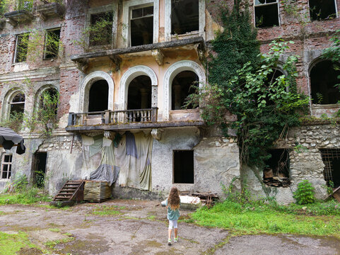 Young girl walking towards a large abandoned building in city of Akarmara, Abkhazia. Dilapidated historical mansion facade with broken windows. Old architecture overgrown with green vines