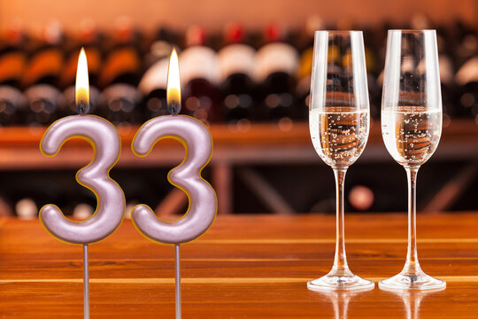 Birthday card, anniversary or celebration of a special day, lit candles forming the number 33 and two champagne glasses; Photo on a background of a bookshelf with blurred wine bottles.
