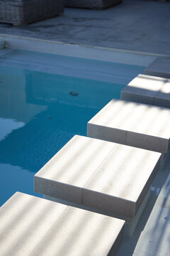 Concrete stepping stones forming diagonal path across clear pool water, casting slatted shadows