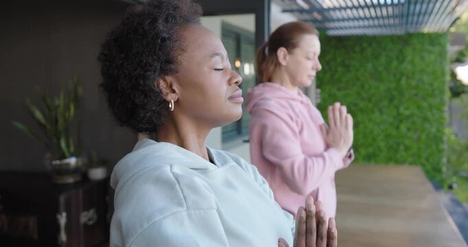 Diverse women friends breathing deeply lifting arms joining palms for calm on patio with green wall