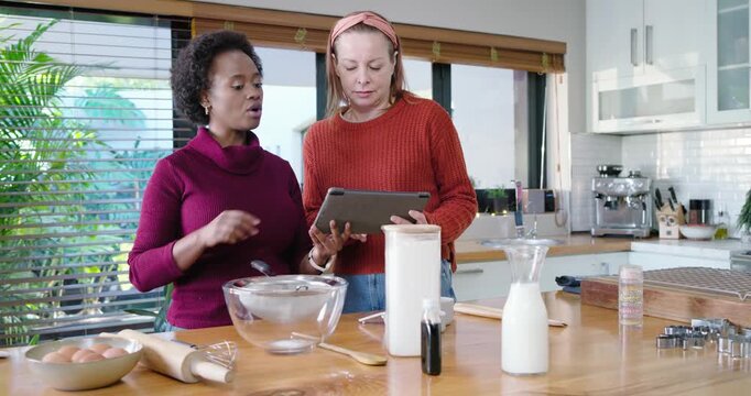 Diverse women reading tablet on kitchen island, alternating holding and measuring eggs for batter