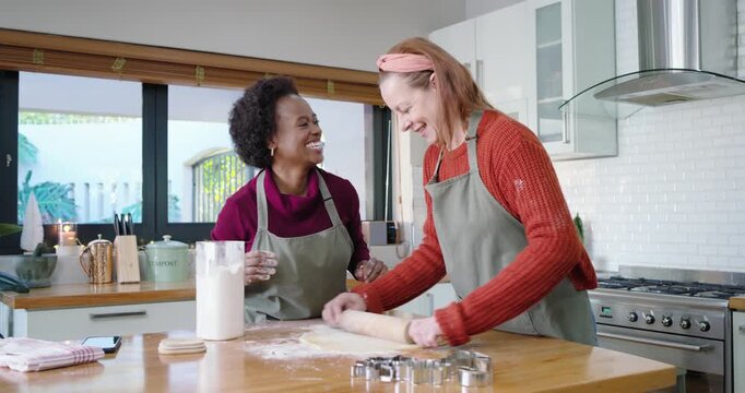 Diverse women at island as left tapping flour right using rolling pin and cutters making cookies