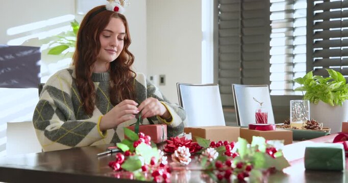 Adult woman sitting at dining table, tying green ribbon around red box, preparing holiday gifts