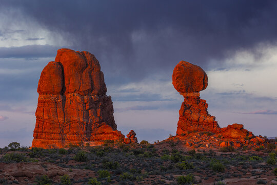 View of the iconic Balanced Rock formation illuminated by golden sunset light against a backdrop of dramatic dark storm clouds in the desert landscape of Moab, Utah, United States.