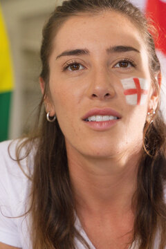 Mid adult woman cheering at stadium, showing right cheek cross paint, white top and hoop earrings