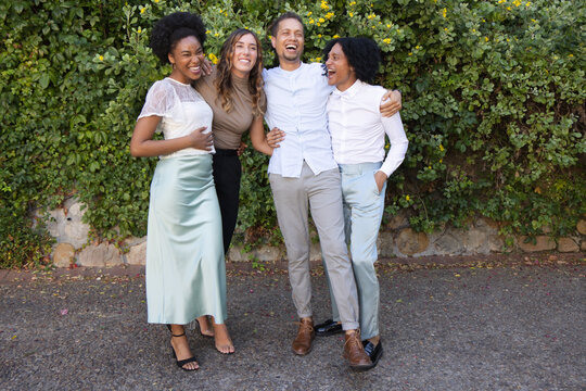 Diverse friends posing and laughing while linking arms on paved walkway by hedge with satin skirt