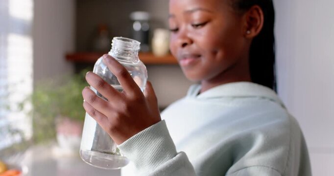 African youth female in hoodie holding bottle checking level tipping and sipping water in kitchen