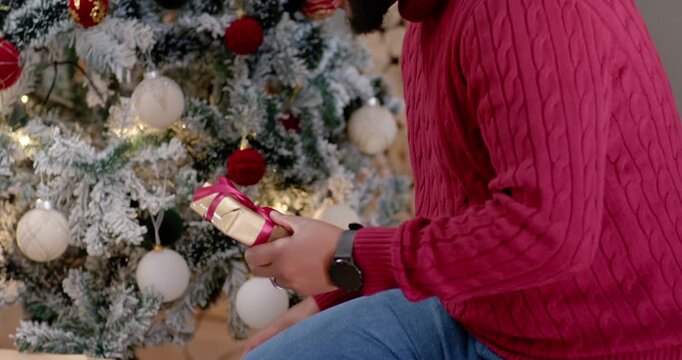 African American father in Santa hat reaching gift by tree with teen daughter leaning, gifting