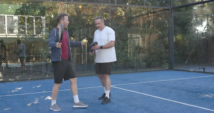 Male coach stepping, showing swing with paddle, ball on padel court teaching senior male trainee