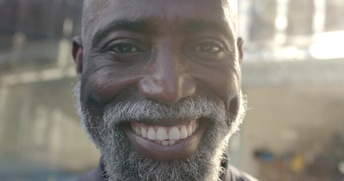 Mature African man smiling facing camera by concrete wall, jacket, eyes shifting sunlight flaring