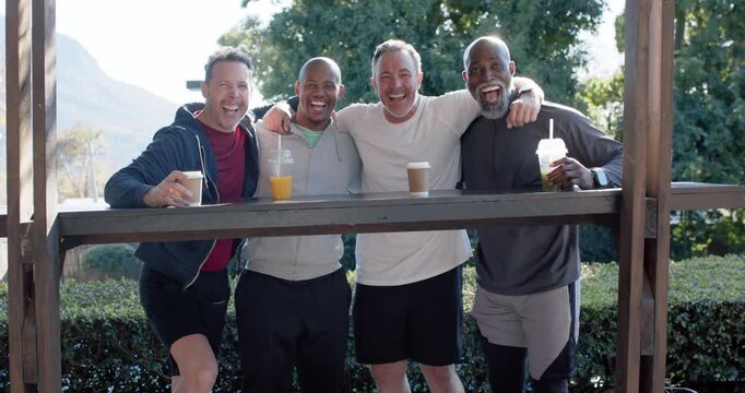 Diverse male friends in sportswear linking arms, laughing, holding coffee at park railing after run