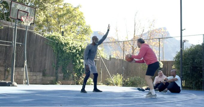 Diverse men at court, defender raising arm, shooter shooting orange ball into hoop to score