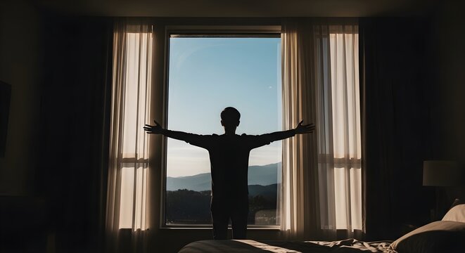 Silhouette of a man standing with arms outstretched in front of a large window overlooking mountains at sunrise