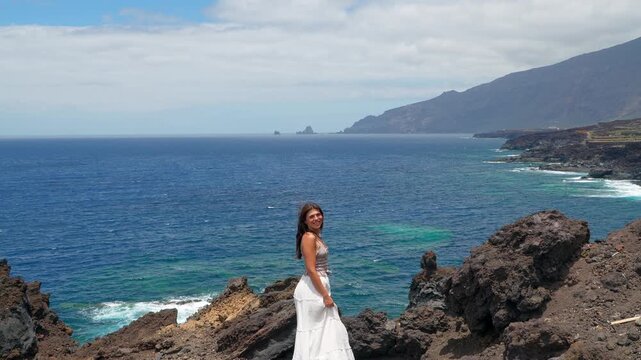 Young woman in white dress turning around on volcanic coastline