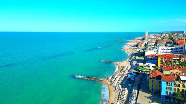Sumptuous aerial push-in truck left pan right arcing shot of the Porto Recanati shoreside, capturing the flowing aquamarine Adriatic Sea waters creating white foam by the sandy beach, with parasols