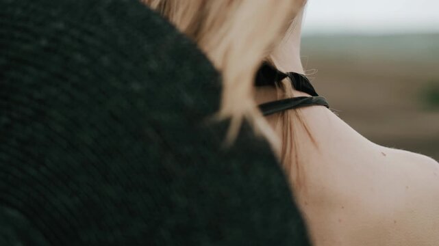 Tight rear view of blonde woman with hair lifted and fanned by wind across neck and upper back, thin black straps and knotted detail visible against muted outdoor landscape and soft sky