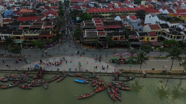 Dense Cluster Of Wooden Boats Moored At Hoi An Quay With Vendors On Shore Crowded Waterfront, Boatmen Arranging Oars, Tourists Gathering For Rides, Colorful Hulls Forming Radial Pattern