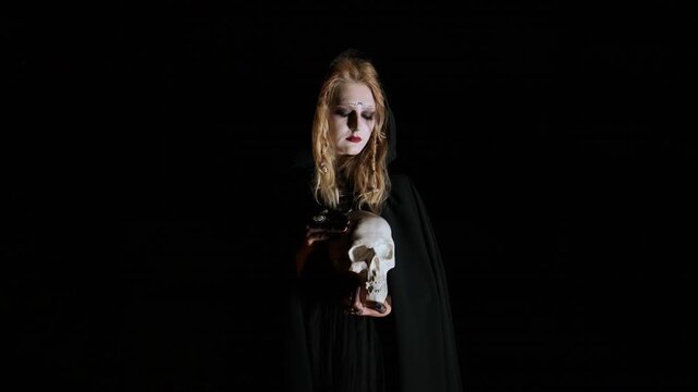 Solitary adult woman in black hooded cloak holds human skull with both hands, head bowed and eyes downward, creating solemn contemplative mood, eerie gothic atmosphere, strong shadows and contrast