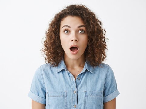 Portrait of a shocked young woman with curly brown hair looking at the camera with wide eyes and open mouth in disbelief, wearing a blue denim shirt against a plain neutral light grey background.