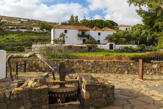 View of the historic white building and traditional stone mill in the foreground, surrounded by lush green vegetation and palm trees in Betancuria, Canary Islands, Spain.