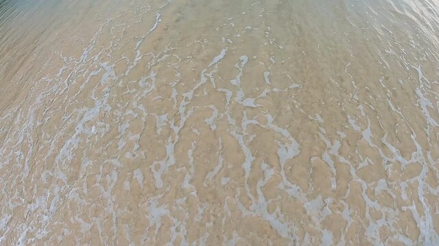 Soft surf spreads across smooth beige sand in high angle coastal view, with pale blue water, thin foam lines, muted tones, and quiet empty mood that suits relaxation, travel, summer, and nature themes