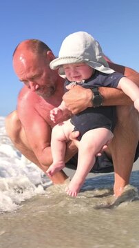 Vertical screen Elderly man with bald head in swim shorts lifts baby in sun hat above the shallow ocean water, surrounded by gentle waves and a clear blue sky, capturing a joyful moment at the beach