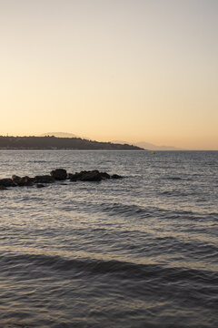 View of the calm Ionian Sea at sunset with dark rocks protruding from the water and distant hills under a soft golden sky in Argassi, Greece.