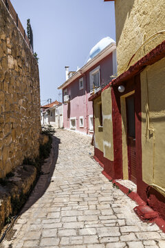 View of a narrow cobblestone street lined with colorful traditional buildings and a rustic stone wall under a clear blue sky in Zakinthos, Greece.