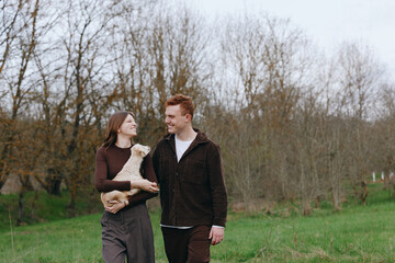 Obraz premium Casual couple outdoors with a goat kid, smiling and holding hands, in a green meadow under soft daylight, a wholesome lifestyle moment that feels natural, warm, intimate, and freely candid.