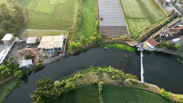 Static aerial drone video of a river with a small dam flowing through a rural village. Surrounding landscape features green agricultural fields, houses, and a road bridge under morning sunlight.