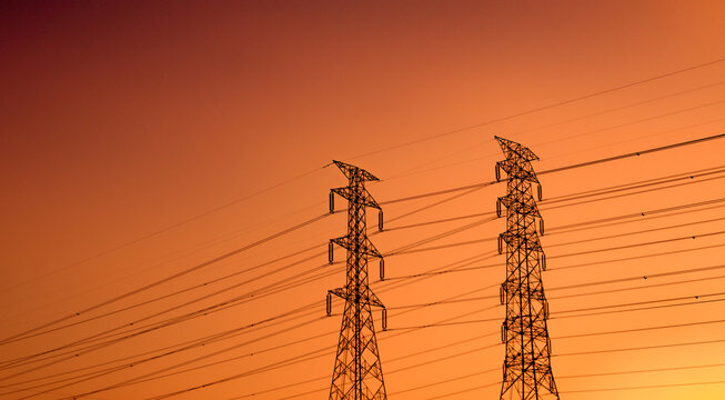 Silhouette of high voltage electricity transmission towers connected by overhead power lines during vibrant orange sunset. National power grid infrastructure, engineering and energy security.
