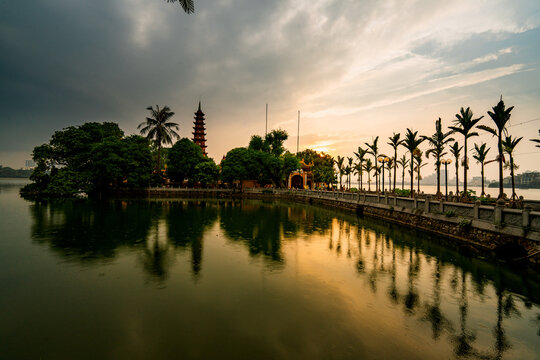 Tay Ho, Vietnam - 16 June 2024: View of Tran Quoc Pagoda at sunset with palm trees lining the stone walkway along the calm waters of West Lake.