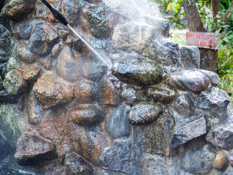 Older man performing cleaning and maintenance work on a stone ornamental water fountain using a pressure washer. The scene shows outdoor maintenance work in a garden surrounded by lush tropical vegeta