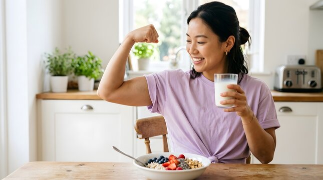 Young happy Asian woman flexing strong arm with a glass of milk at a healthy breakfast in the kitchen