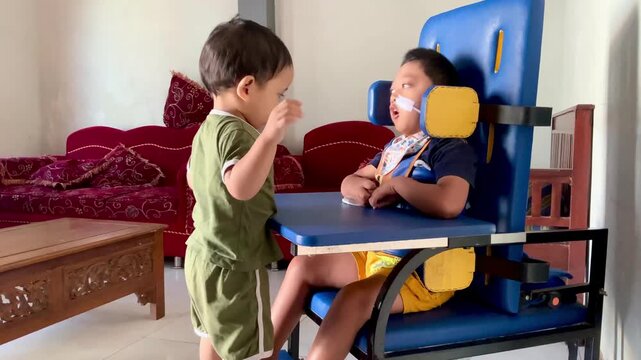A video clip showing a toodler interacting with their brother who is seated in a specialized adaptive chair within a home setting, highlighting family connection and daily life.