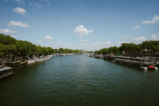 Paris, Ile-de-France, France - 06 May 2022: View of the Seine River flowing through Paris with tour boats moored along the tree-lined banks under a bright blue sky with scattered clouds.